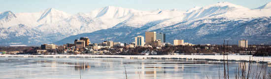 Stunning winter city skyline reflecting in a river, surrounded by snow-capped mountains.